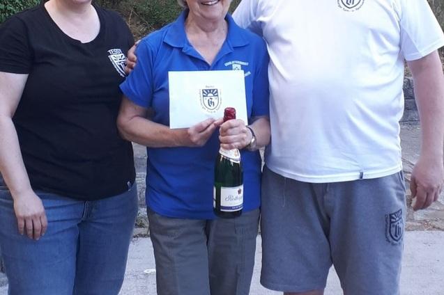Drei Personen stehen zusammen, eine hält eine Flasche Sekt und eine Karte, vor einer grünen Landschaft im Hintergrund.