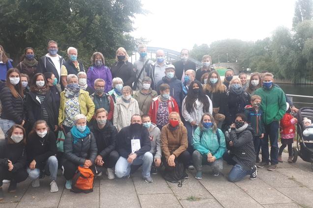 Gruppenzusammenkunft von 40 Personen am Ufer eines Gewässers, alle tragen Atemschutzmasken und unterschiedliche Oberbekleidung.