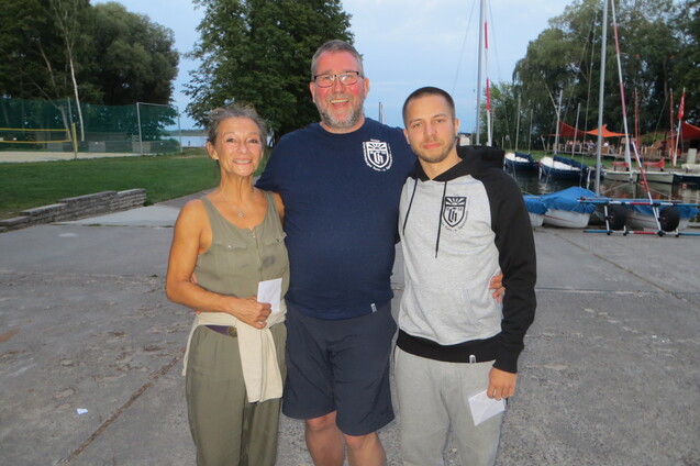Drei Personen stehen lächelnd an einem Hafen, umgeben von Bäumen und Booten im Hintergrund.