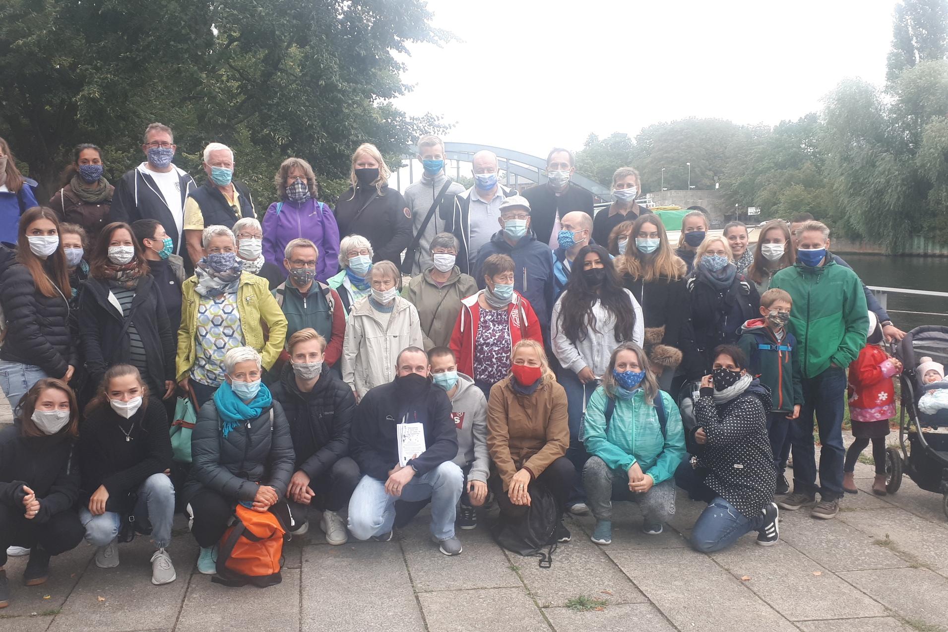 Gruppenzusammenkunft von 40 Personen am Ufer eines Gewässers, alle tragen Atemschutzmasken und unterschiedliche Oberbekleidung.