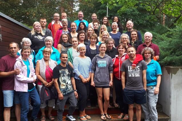 Gruppenfoto von etwa 40 Personen, die im Freien vor einer Holzwand stehen, umgeben von Bäumen und Pflanzen.