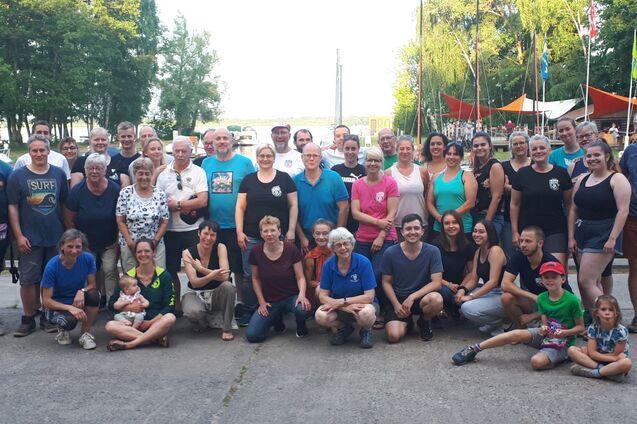 Gruppenfoto von etwa 50 Personen am Wasser, umgeben von Bäumen und Segelbooten, in fröhlicher Atmosphäre.