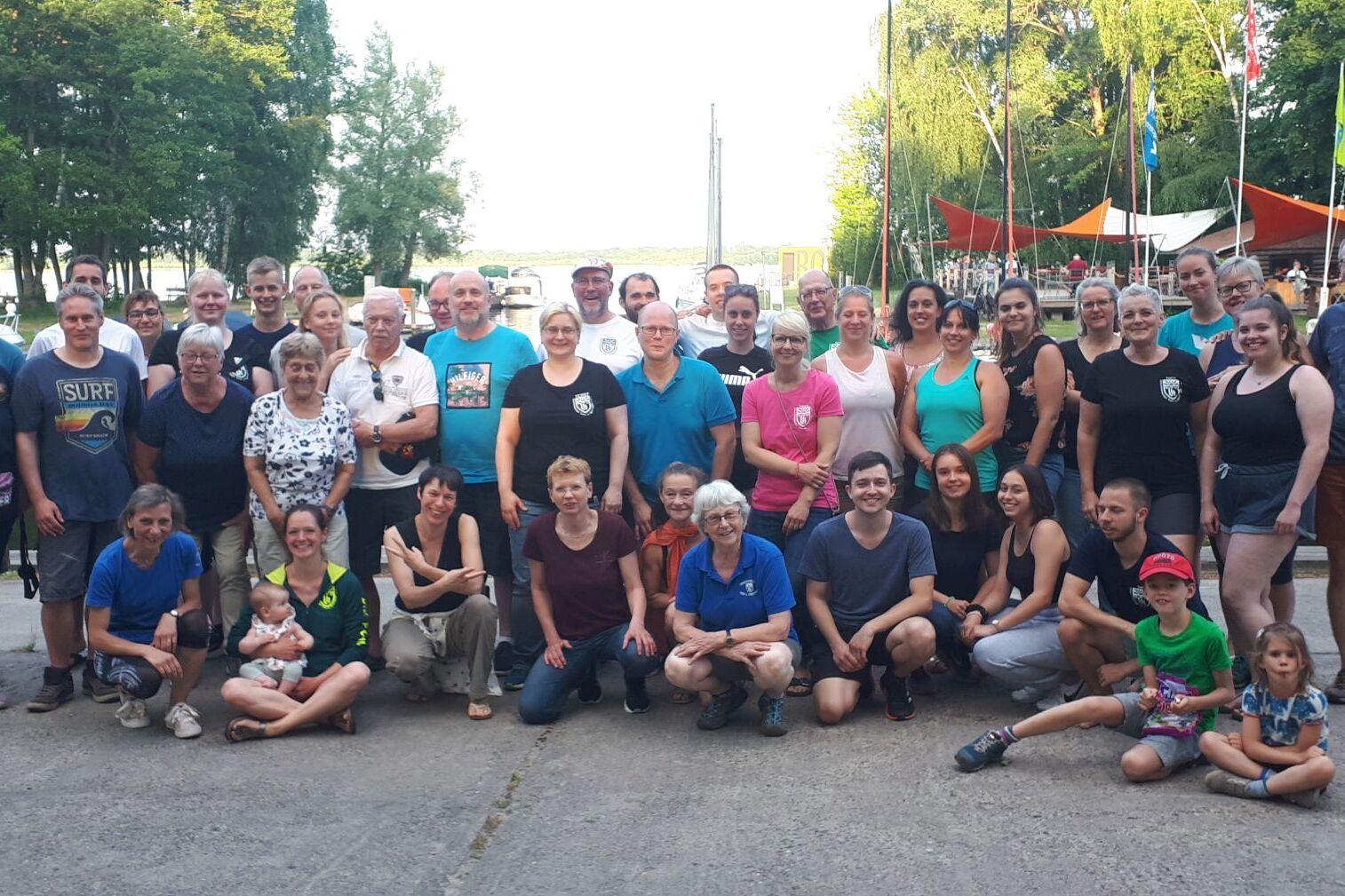 Gruppenfoto von etwa 50 Personen am Wasser, umgeben von Bäumen und Segelbooten, in fröhlicher Atmosphäre.