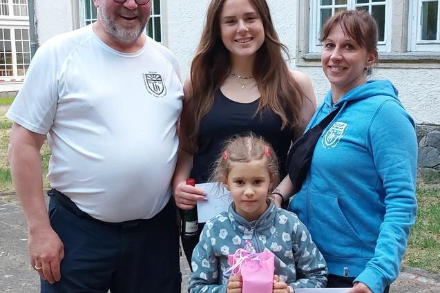 Vier Personen stehen lächelnd zusammen im Freien, eine Frau hält ein pinkes Geschenk, alle zeigen Dokumente.