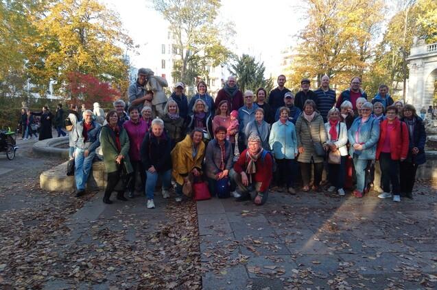 Gruppe von etwa 40 Personen posiert im Park vor einem Denkmal, umgeben von buntem Herbstlaub.