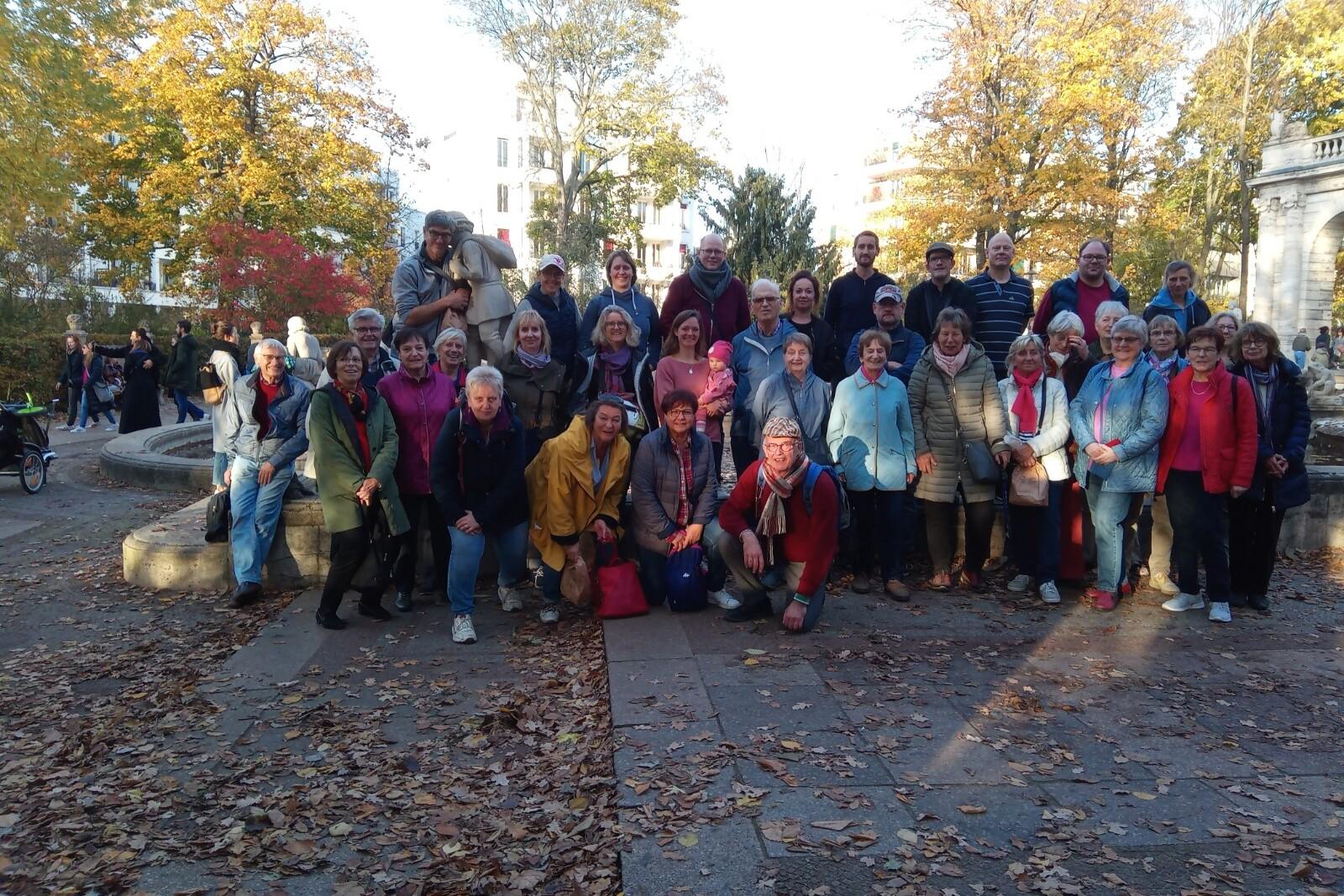 Gruppe von etwa 40 Personen posiert im Park vor einem Denkmal, umgeben von buntem Herbstlaub.