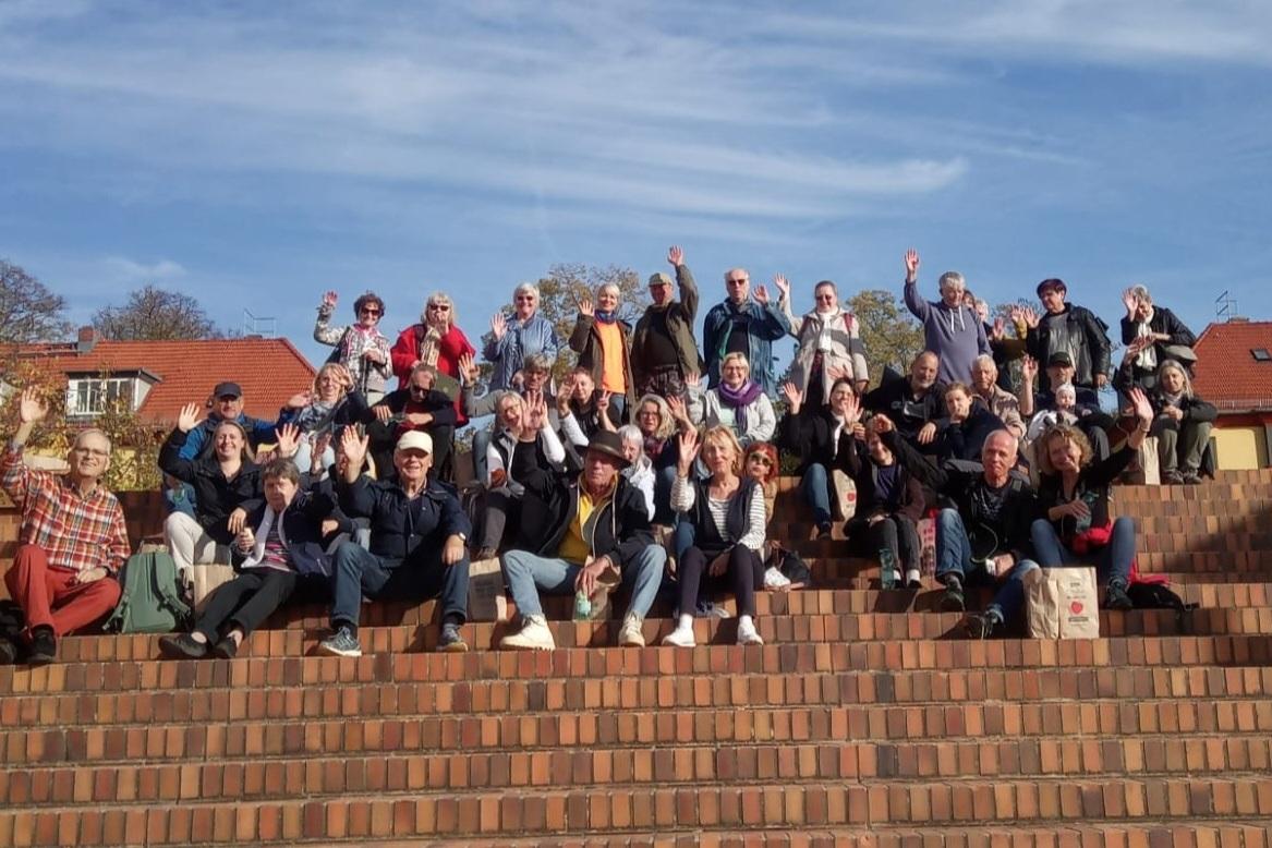 Gruppenszene mit 43 Personen auf Stufen, die in die Kamera winken, bei klarem Wetter und blauem Himmel.