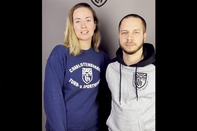 Zwei Personen stehen vor einem grauen Hintergrund, beide tragen Sweatshirts mit Vereinslogos.
