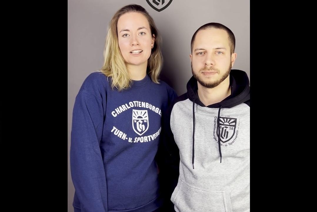 Zwei Personen stehen vor einem grauen Hintergrund, beide tragen Sweatshirts mit Vereinslogos.