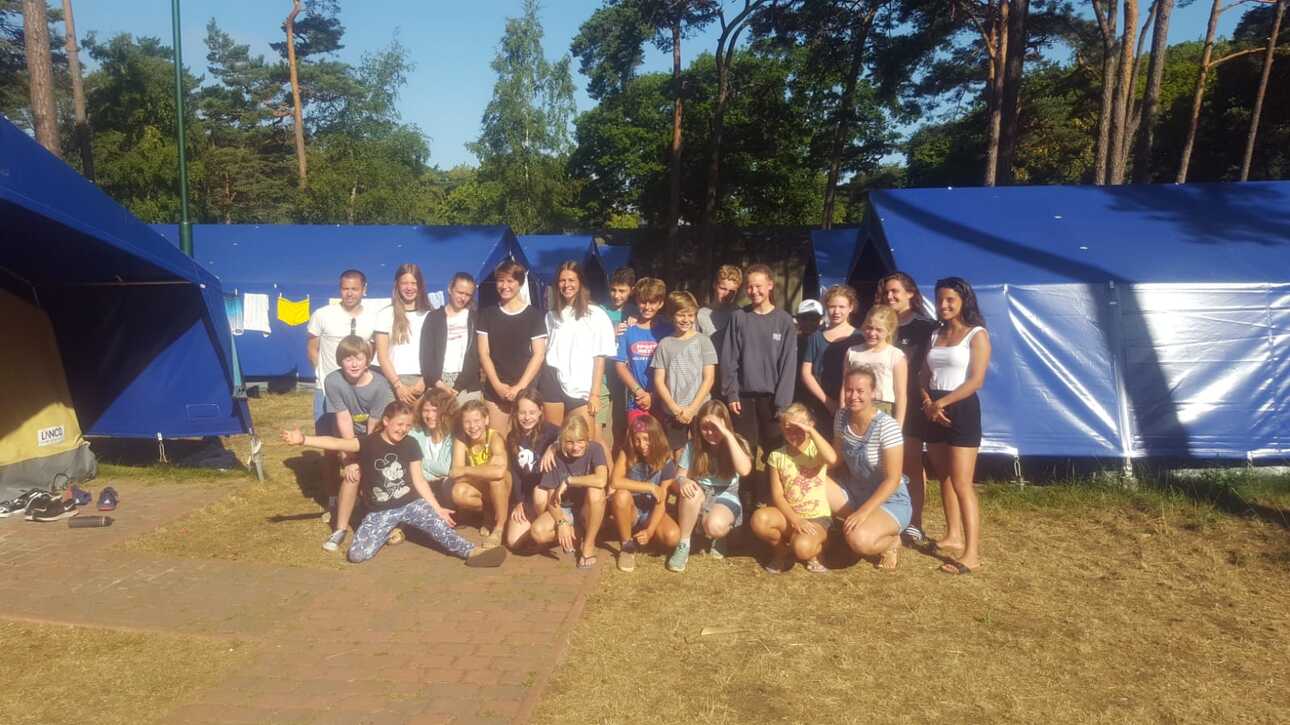 Gruppenfoto von Jugendlichen und Betreuern vor blauen Zelten in einer sonnigen, bewaldeten Umgebung.