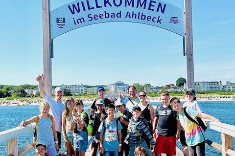Gruppe von Kindern und Erwachsenen posiert unter dem Willkommensschild am Seebad Ahlbeck auf einem Holzsteg.