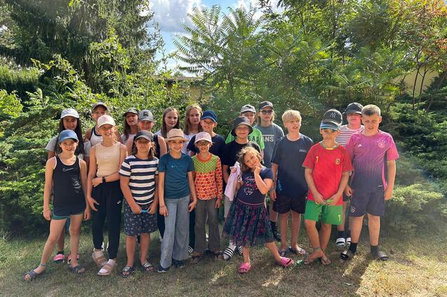 Gruppe von 20 Kindern steht vor Büschen, die meisten tragen Hüte, sonnige Stimmung, verschiedene Kleidungsstile.