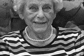 Ältere Frau mit grauen Haaren, gestreiftem Shirt und Perlenkette, lächelt auf einem gepolsterten Sofa.