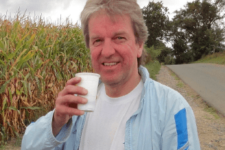 Mann mit grauen Haaren hält einen weißen Becher, steht an einem landwirtschaftlichen Weg neben einem Maisfeld.