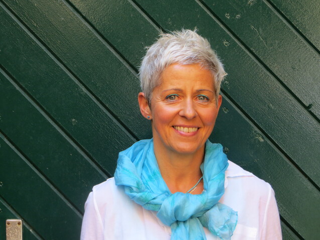Porträt einer Frau mit kurzem, grauem Haar und blauer Schal vor einer grünen Holzfläche. Sie lächelt freundlich.