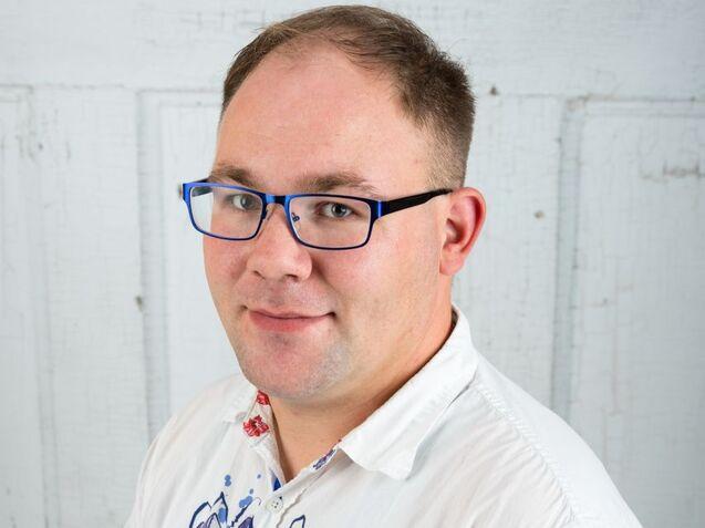 Porträt eines Mannes mit kurzen Haaren und blauen Brillengestell, der ein gemustertes Hemd trägt, vor einer weißen Wand.