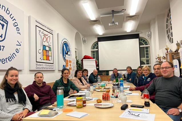 Teilnehmer an einem runden Tisch in einem Konferenzraum, umgeben von Präsentationsmaterialien und Getränken.