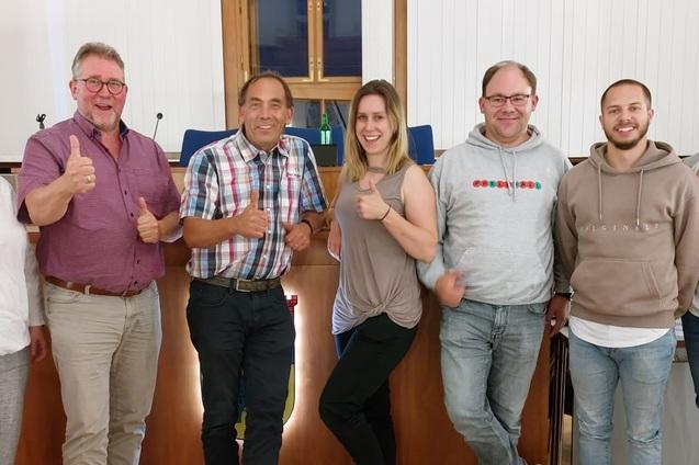 Gruppe von sieben Personen mit Daumen hoch vor einem Holztisch in einem Raum mit Fenstern.