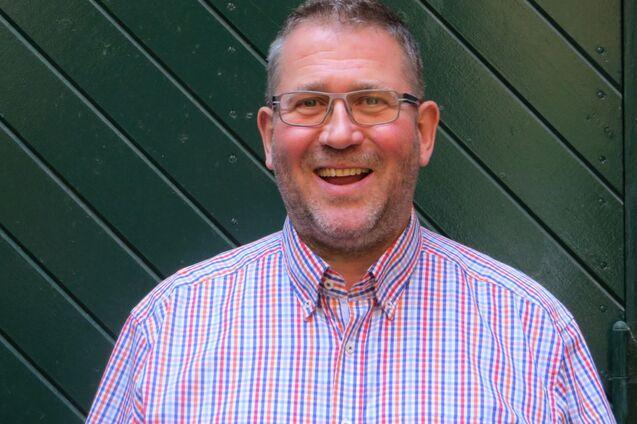 Lächelnder Mann mit Brille, trägt ein karriertes Hemd, vor einer grünen Holzwand.