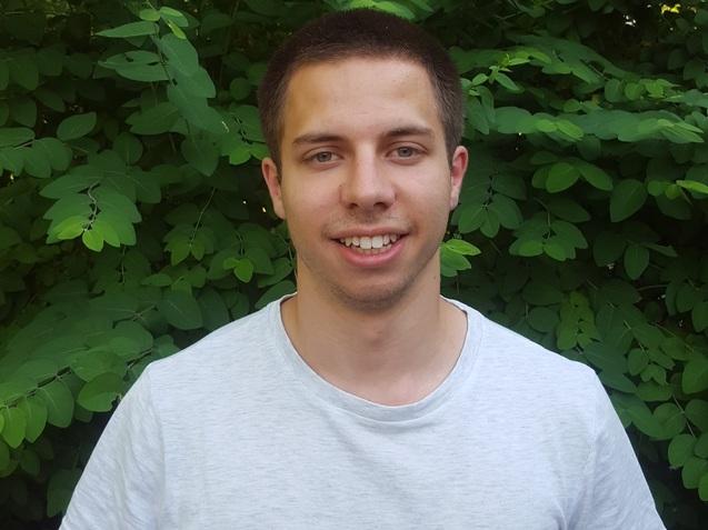 Junger Mann in einem grauen T-Shirt steht vor einem Hintergrund aus grünen Pflanzenblättern.