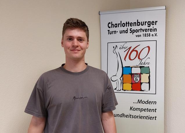 Junger Mann mit grauem T-Shirt steht vor einem Banner des Charlottenburger Turn- und Sportvereins.
