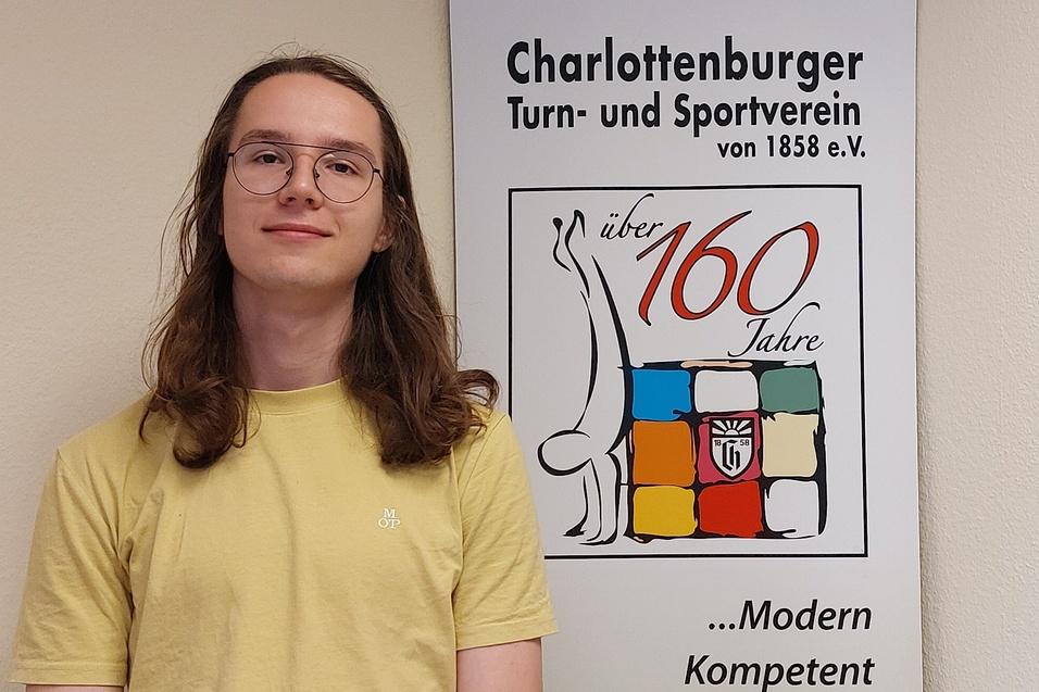 Junger Mann mit langen, lockigen Haaren steht vor einem Banner des Charlottenburger Turn- und Sportvereins.