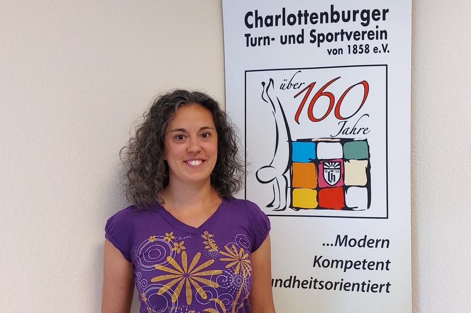 Frau mit lockigem Haar lächelt vor einem Banner des Charlottenburger Turn- und Sportvereins, das 160 Jahre älter ist.